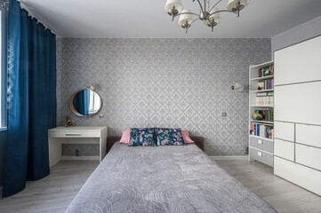 A tidy bedroom with a single bed, patterned wallpaper, blue curtains, a chandelier, white desk,...