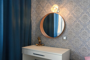 A stylish corner with a round wooden mirror, wall lamp, white dresser, golden figurine, patterned...