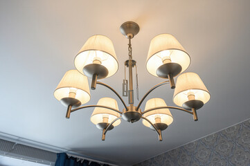 A modern ceiling chandelier with eight cream lampshades, metallic frame, suspended by chain; emits...