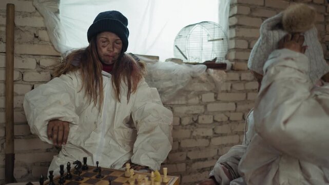 Dirty child scratches wool hat while another makes chess move and taps clock inside cold brick-walled room, showing survival mood with broken birdcage and plastic covered window in background