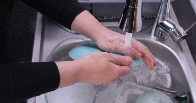 Close-up of rinsing a plate covered in foam under running water in a kitchen sink. Dish washing, cleanliness and detergent effectiveness concept. Slow motion