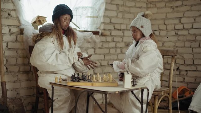 Two orphan girls in worn white winter clothing play chess in abandoned indoor space. Younger sister passes black chess piece to elder while both sit focused in cold rustic room
