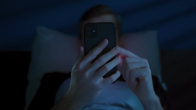 Close-up of unrecognizable man lying in bed at night, holding mobile phone in front of his face while consuming social media content. Gadget addiction, excessive night screen time. Smartphone overuse 