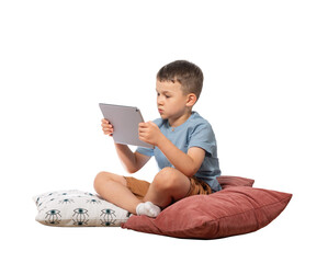 The boy looks at the tablet computer screen while sitting on pillows. Isolated on white background.