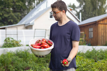 A man in a dark T-shirt is standing in front of his garden with a plastic basin filled with red peppers. A young man with glasses is holding out a red pepper.