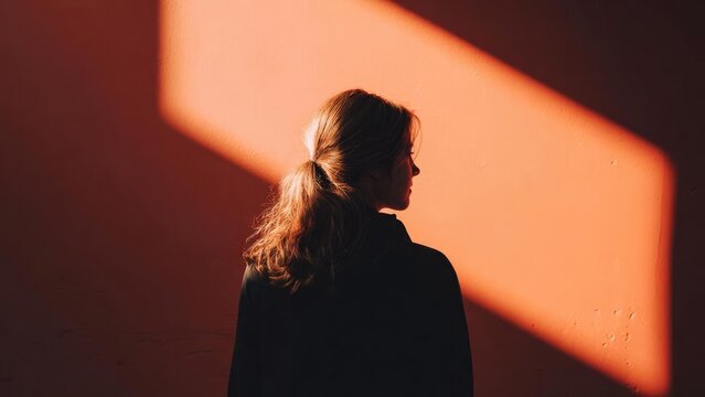 Person in profile with a ponytail stands against a warm orange wall, lit by a bold diagonal shadow. Concept Profile portrait, Ponytail, Warm orange wall, Diagonal shadow, High-contrast lighting