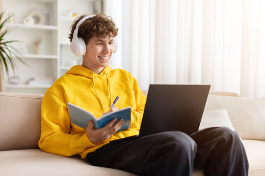 Smiling teenage guy wearing yellow hoodie and headphones, studying at home with laptop and notebook, writing notes, enjoying online learning, focus, and comfortable modern lifestyle indoors