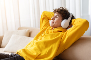 Relaxed teenage boy in yellow hoodie leaning back on couch at home, listening to music with headphones, eyes closed, enjoying calm leisure time and comfortable modern lifestyle indoors © Anastasiya