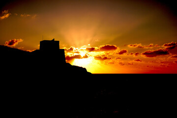 Fototapeta premium Dramatic silhouette of ancient fortress ruins on hilltop against vibrant orange and red sunset sky, Malta historic landscape at golden hour dusk