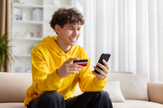 Teenage guy smiling while checking smartphone and holding credit card, sitting on sofa at home, wearing yellow hoodie, enjoying online shopping and digital lifestyle indoors