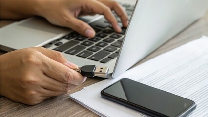 A person using a laptop with a USB drive in hand, alongside a smartphone and documents on a wooden desk.