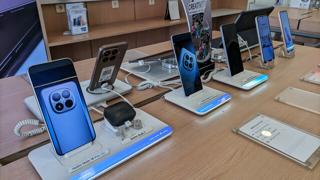 A smartphone display case in an electronics store, several Redmi phones are displayed on a wooden table with security stands and illuminated screens, showcasing the latest product features.