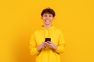 Fototapeta premium Teenage guy holding smartphone and smiling on yellow studio background, modern youth communication and technology lifestyle concept with clean copy space