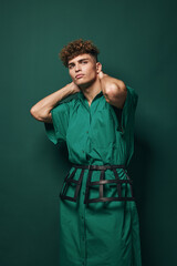 Young man with curly hair wearing green dress and black belt posing with hands behind neck against...