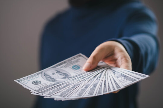 Hand holding and giving cash money in US dollar bills, representing financial transaction, cash payment, income, profit, salary, business finance, investment return, and economic exchange concept.