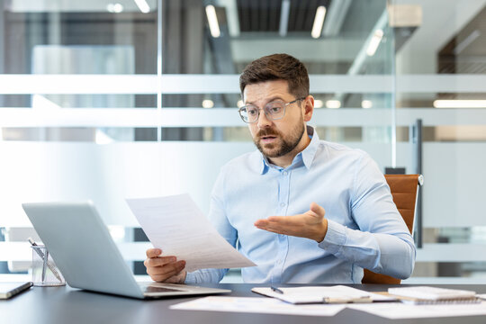 Confused businessman at office desk reading stressful financial reports and paperwork, showing concern and frustration while analyzing documents and laptop during a tense workday