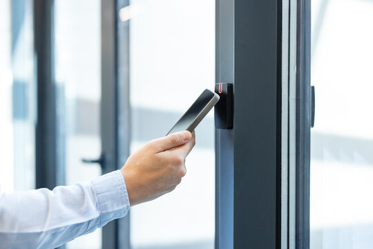 Person's hand holding a smartphone to a contactless card reader on a door frame, performing an access control operation for security in an office or commercial building