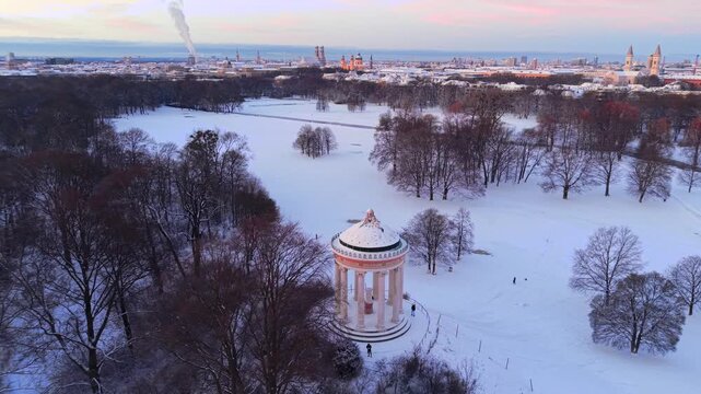 Breathtaking winter sunrise over Munich, Germany. Aerial drone view of Monopteros in English Garden with the historic old town center and Frauenkirche towers visible in the snowy distance