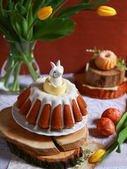 Traditional Easter cake with ceramic bunny on wooden stand and yellow tulips in vase. Concept of Easter celebration, festive food styling background for banner, card, retail marketing.