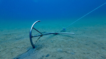 Fototapeta premium Metal anchor lying on a sandy seabed underwater. Minimal underwater scene shows a boat anchor attached to a rope resting on the ocean floor in clear blue water.