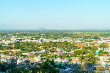 High angle view of a small Thai city with colorful rooftops, a traditional Buddhist temple, and a tall communication tower. Scenic suburban landscape with green trees and mountains under a blue sky.