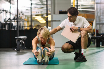 Caucasian young adult woman performing seated forward stretch on exercise mat, while Caucasian young adult man physical therapist supervising and providing guidance in rehabilitation setting