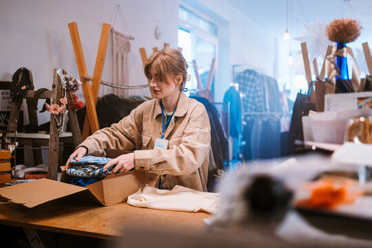 Young Woman Of Caucasian Descent Carefully Packing Clothes Into Boxes At Quaint Store For Future Use