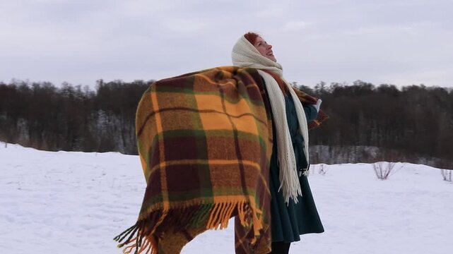 A woman on a snowy mountain opens a warm blanket, breathing in the frosty air. Winter freedom, the peace of nature, a feeling of lightness and inner harmony.