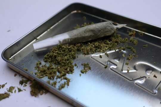 Hand rolled marijuana joint resting on metal rolling tray surrounded by ground cannabis herb, medical cbd and recreational thc consumption concept