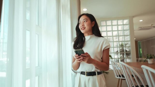 Cheerful Asian woman smiling while texting on her phone. She is standing by a window in a modern, bright living room and looks thoughtful