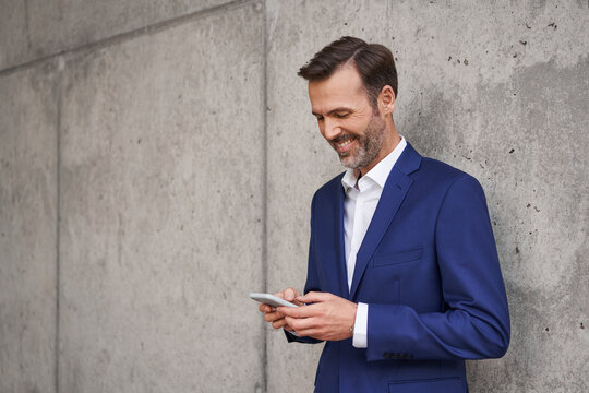 Happy businessman using mobile phone leaning against concrete wall