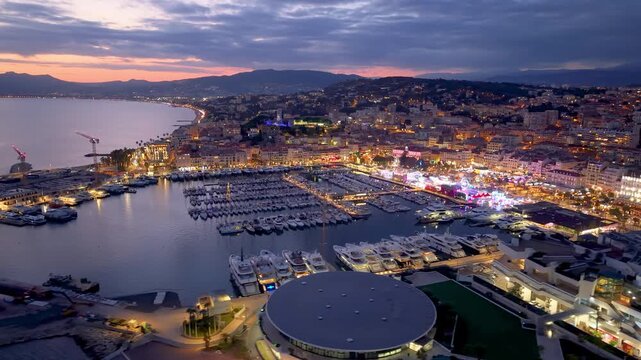 Aerial Footage Showcases Vibrant Waterfront Illumination Against City And Mountain Skyline Backdrop. Cannes. France