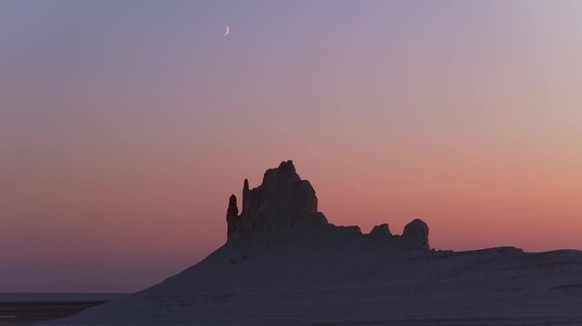 Aerial view of a stark rock formation silhouetted against a gradient sky with a crescent moon, creating a serene desert landscape, Aktau, Mangystau Region, Kazakhstan.