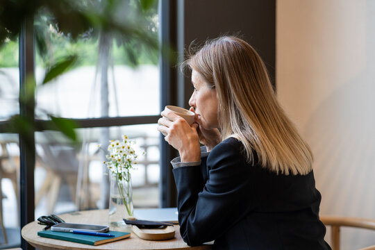 Pensive businesswoman freelancer enjoying aromatic coffee in stylish restaurant on lunch break during workday. Confident female drinking from cup looking outside comfortable sitting at table in cafe.