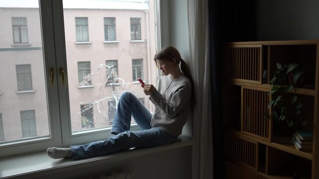 Surprised interested woman scrolling social media with smartphone rest by window at home, smiling looking at screen. Happy pleased young female online chatting with phone relaxed sitting on windowsill