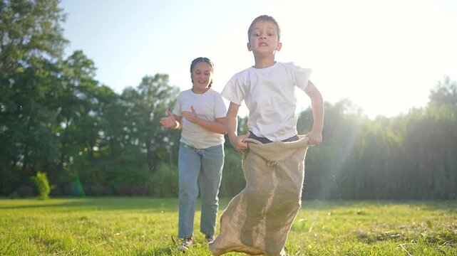 Boy jumps in burlap sack during outdoor race with mother watching. Child plays sack race game in sunny park. Kid jumping in sack on grass. Mother cheers boy during outdoor sack race activity.