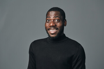 Fototapeta premium African man with dark skin wearing black turtleneck sweater posing with surprised expression in creative conceptual visual style on gray background. Studio portrait of young male model.