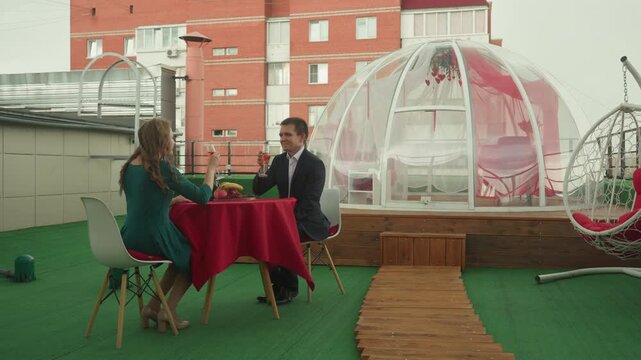 rooftop igloo dinner with white couple, intimate bubble dome on rooftop, red tablecloth and floral centerpiece, woman in green dress pours champagne, man in suit raises glass, clinking and laughter,