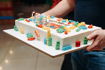 large white birthday cake in adult man hands for his child, son or daughter, cake decorated with edible lego details, lego-form cake, celebrating happy birthday for kids © Anton Pentegov