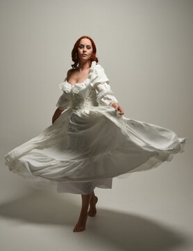 full length portrait redhead female model wearing historical fantasy wedding dress, fairytale ball gown.  isolated figure dancing with flowing fabric on white studio background