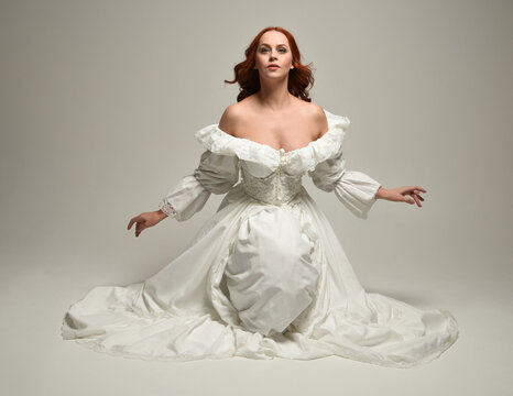 full length portrait beautiful female model wearing historical white fantasy wedding dress, fairytale ball gown. isolated figure sitting pose, hands gesturing  on white studio