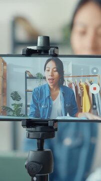 Vertical Screen : Young Asia lady fashion designer using mobile phone receiving purchase order and showing clothes recording video live streaming online at shop. Small business owner, market delivery.