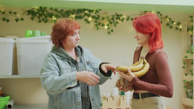 white women hesitant banana exchange in pantry older volunteer appears hesitant while younger customer watches, ambient plants and fairy lights create gentle mood, cluttered shelves with jars