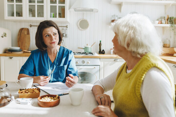 Home visit of female nurse in blue uniform for elderly woman with health issues, social worker collecting complaints before giving medical aid and support both sitting at kitchen table
