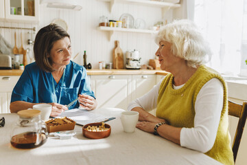 Two people in kitchen, senior lady getting advice and consultation from nurse volunteer at home, healthcare worker in blue uniform collecting complaints and noting down into list before give support