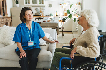 Side view of female kinesiotherapist expert sitting on sofa in blue uniform home visiting her old disabled lady in wheelchair helping to go through rehabilitation treatment after brain stroke