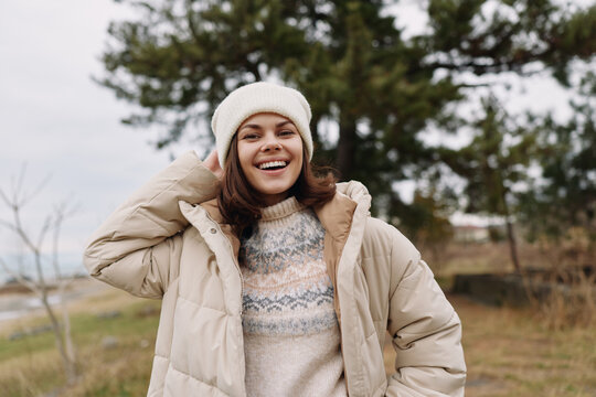 Young woman posing outdoors in a puffer jacket and knit sweater, smiling under a white beanie in chilly weather, enjoying a relaxed moment in a park with muted autumn tones and soft light