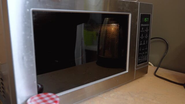 Close up view of a person's hand placing a glass inside a modern stainless steel microwave oven, closing the door and starting the timer to heat up the contents on a kitchen counter