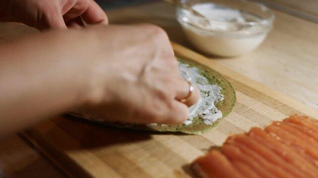 Laying salmon slices as filling on green pancake anointed with creamy cheese on a wooden board