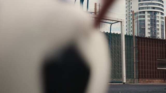 male goalkeeper stands beside goalpost on wet urban pitch, controlling ball during solitary training drill, blue windbreaker fluttering, chainlink fence and highrise skyline in background, rain